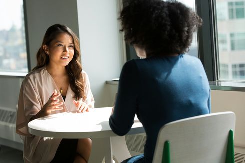 Two women in a bright room; one speaks, smiling. They sit at a round white table.