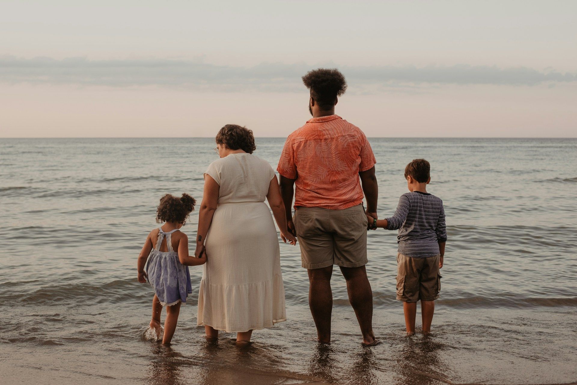 Family of four holding hands at the beach, looking out at the water. Sunset colors.