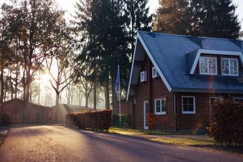 A cabin home with a blue roof sits on a road, trees and sunlight in the background.