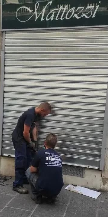 Due operai sono accovacciati davanti alla saracinesca metallica chiusa di un negozio, sotto un'insegna con la scritta 