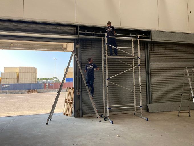 Operai su un'impalcatura riparano l'ingresso di un magazzino sotto una saracinesca metallica parzialmente aperta.