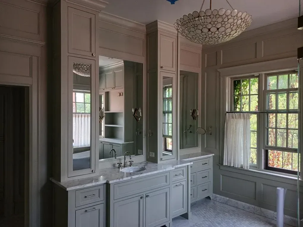 Elegant gray bathroom with double vanity, large mirrors, chandelier, and sunlit windows.