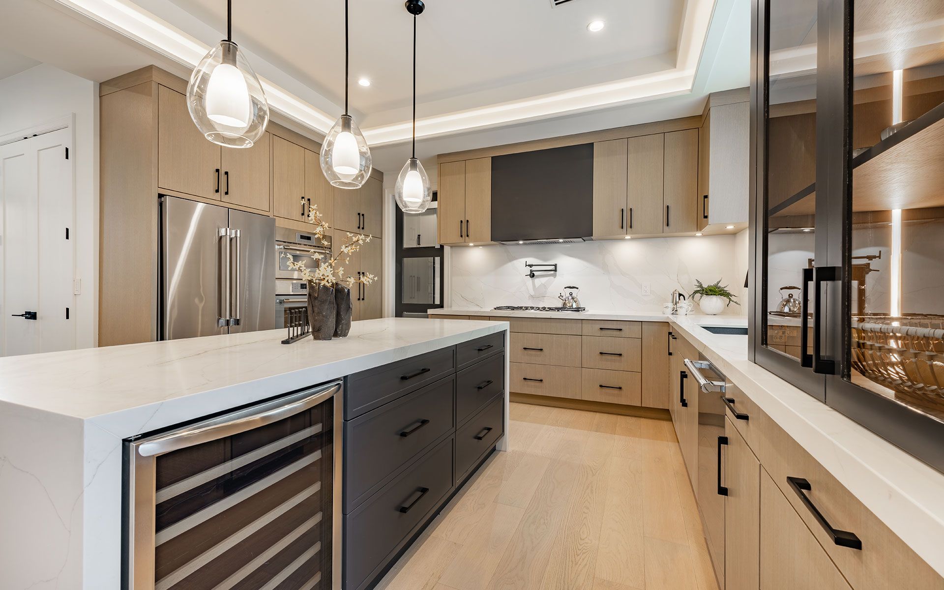 Modern kitchen with light wood cabinets, dark island, stainless steel appliances, and pendant lights.