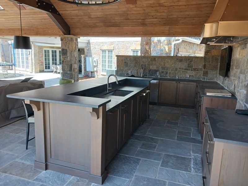 Outdoor kitchen with dark countertops, cabinets, stone walls, and a large sink.