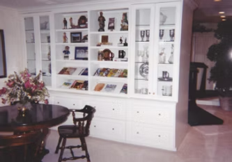 White built-in cabinet with glass doors and open shelving, displaying objects and magazines, next to a table and chair.