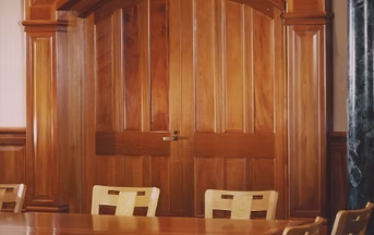 Wooden double doors with arched top and wood paneling in a room with light wood chairs and a table.