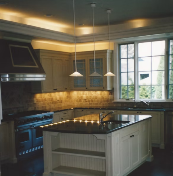 Cream-colored kitchen with island and pendant lights. Dark countertops, stainless steel appliances, and large window.