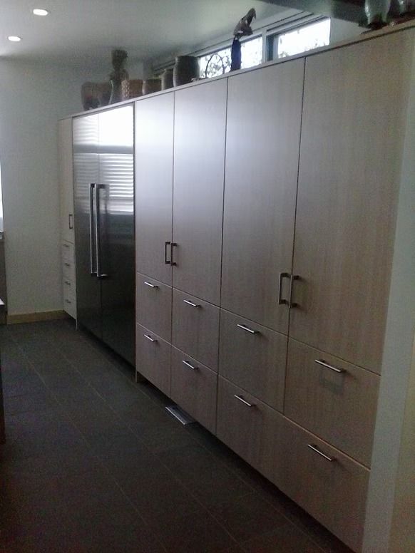 Tall, light-colored kitchen cabinets and stainless steel refrigerator against a white wall and dark floor.