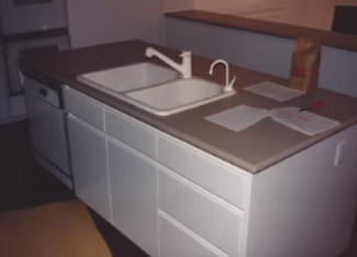 Kitchen island with double sink, faucet, white cabinets, and dishwasher; brown countertop; brown paper bag.