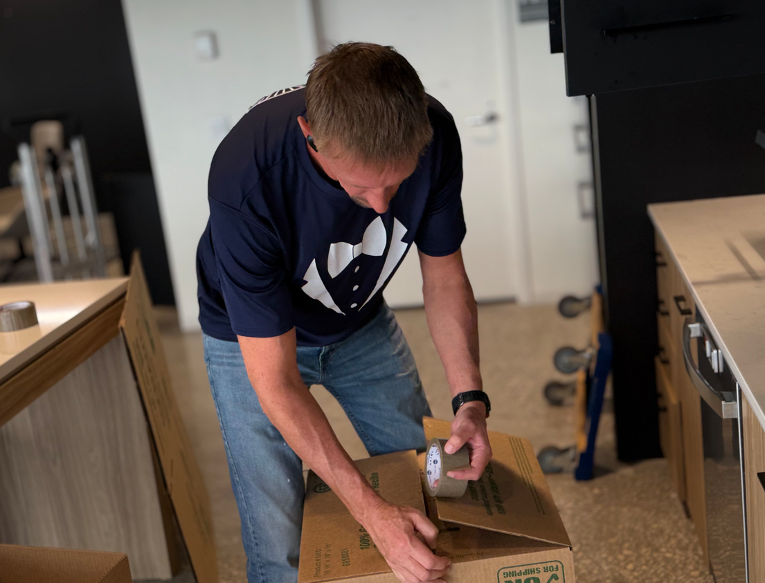 Man in blue shirt and jeans taping a cardboard box in a room with light-colored cabinets.