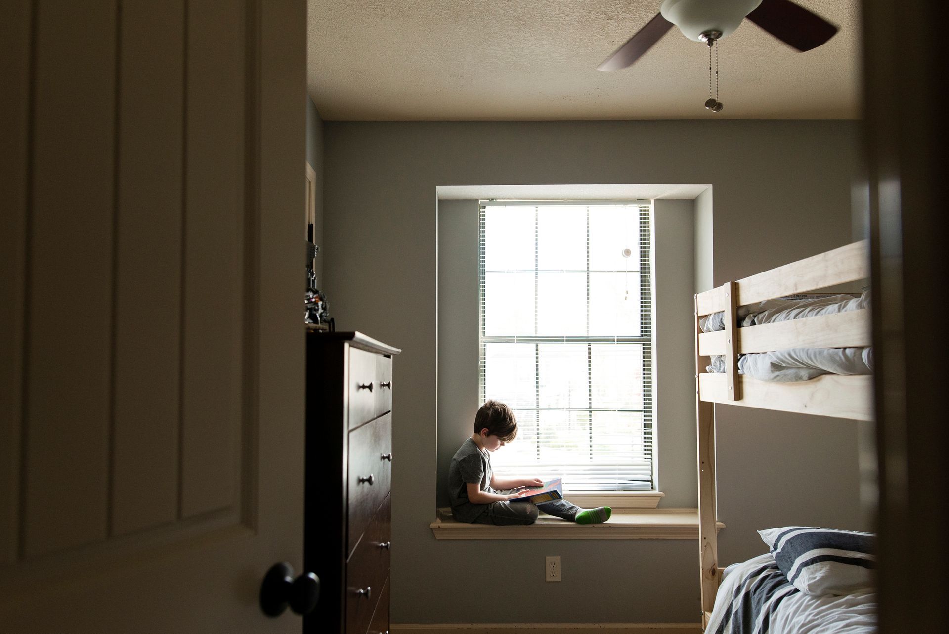 Un bambino legge vicino a una finestra in una camera da letto con un letto a castello e una cassettiera.
