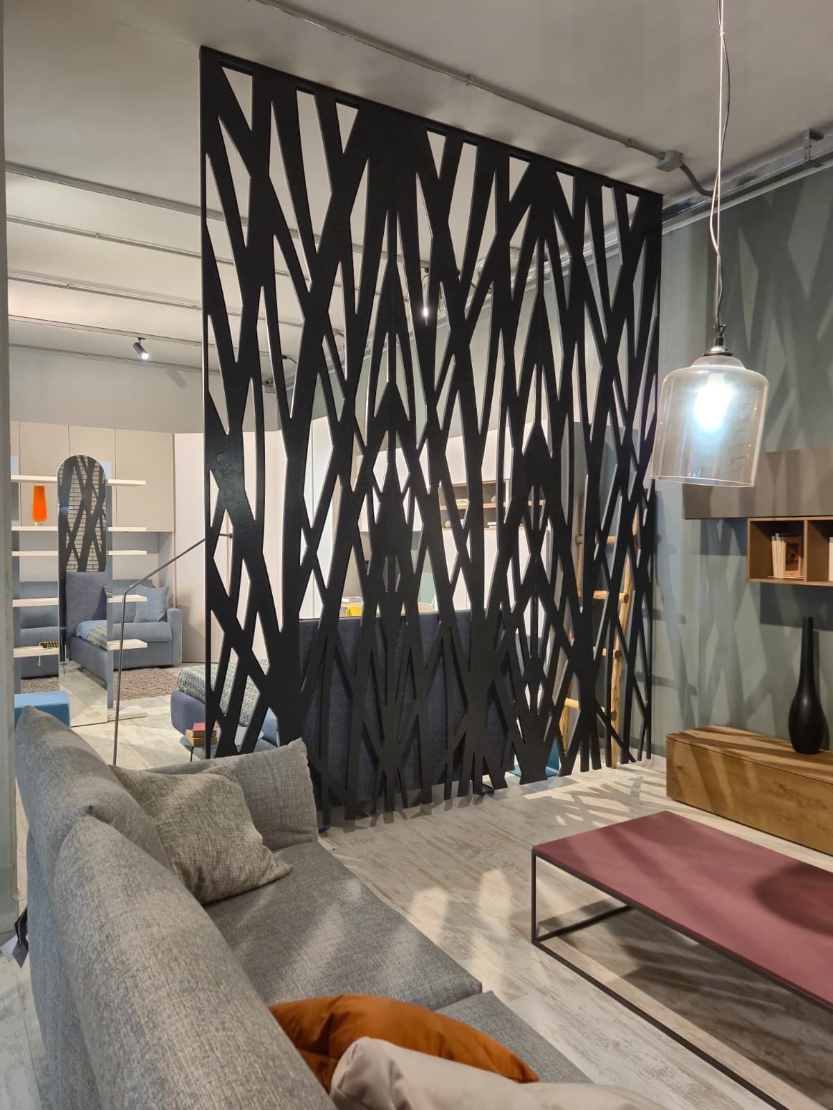 Un separé geometrico nero separa lo spazio abitativo con divano grigio, elementi in legno e illuminazione moderna.