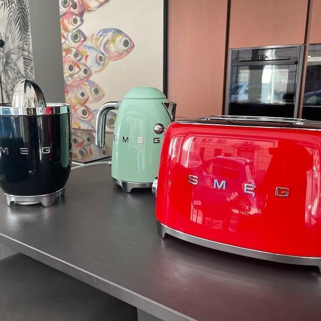 Spremiagrumi nero, bollitore alla menta e tostapane rosso su un piano di lavoro con opere d'arte ed elettrodomestici sullo sfondo.
