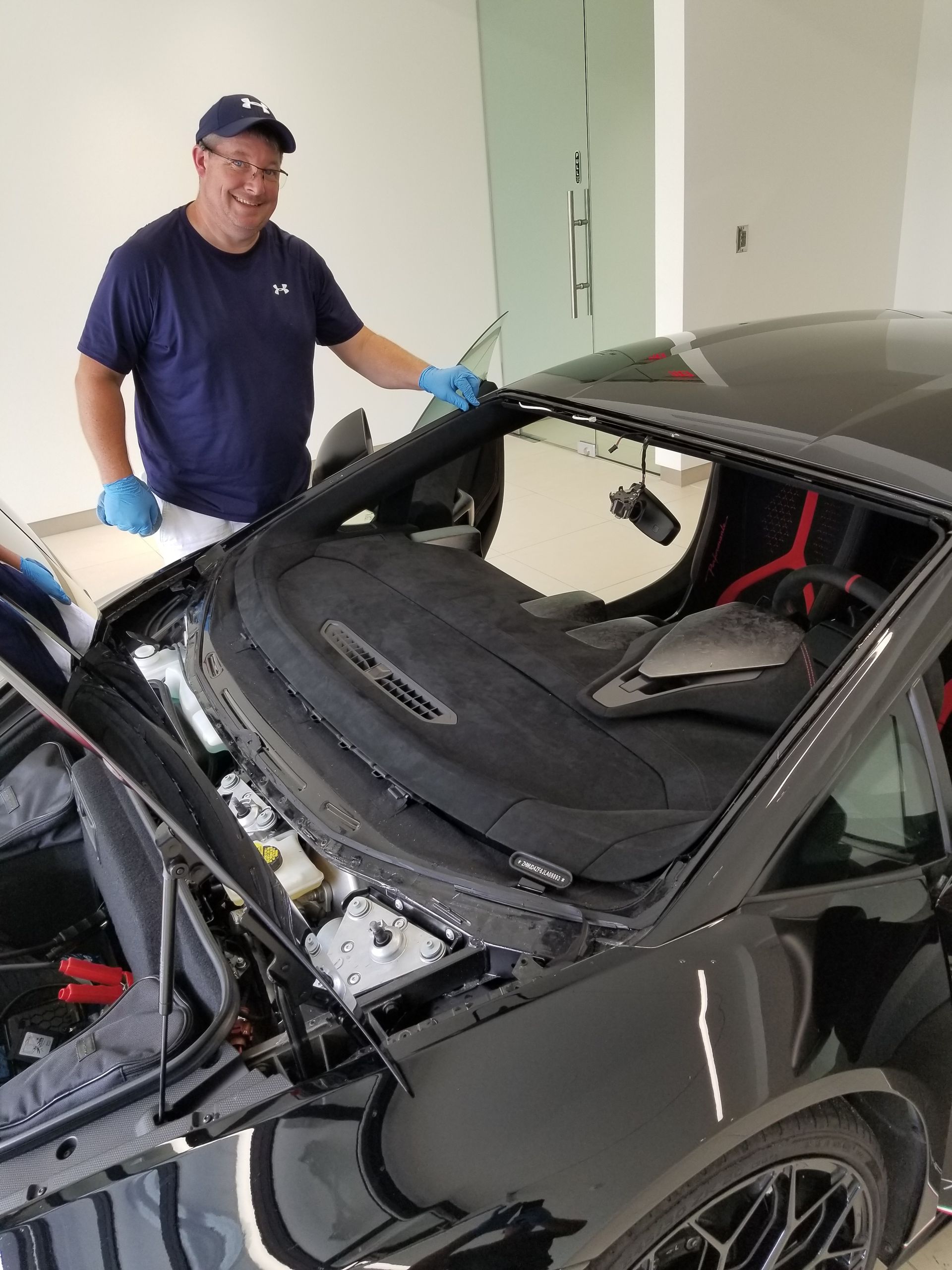 Man in a blue shirt and cap beside a damaged black sports car, possibly in a repair shop.