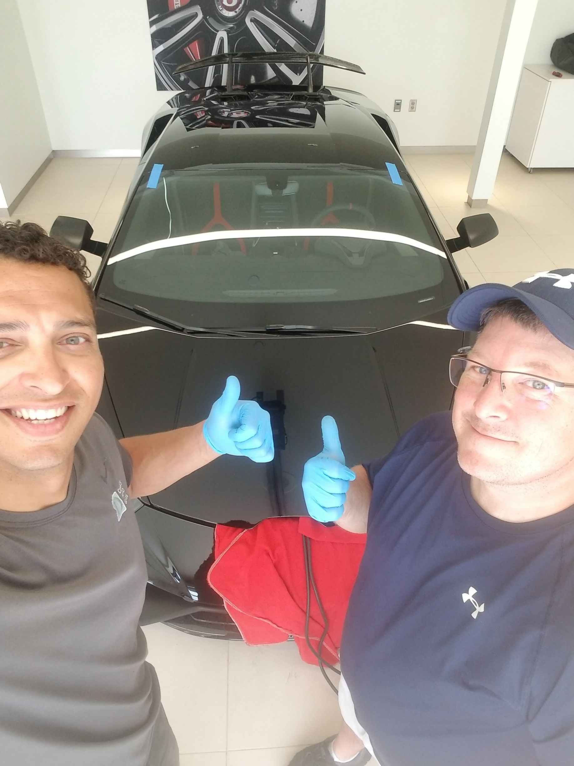 Two men give thumbs up next to a black sports car in a showroom.
