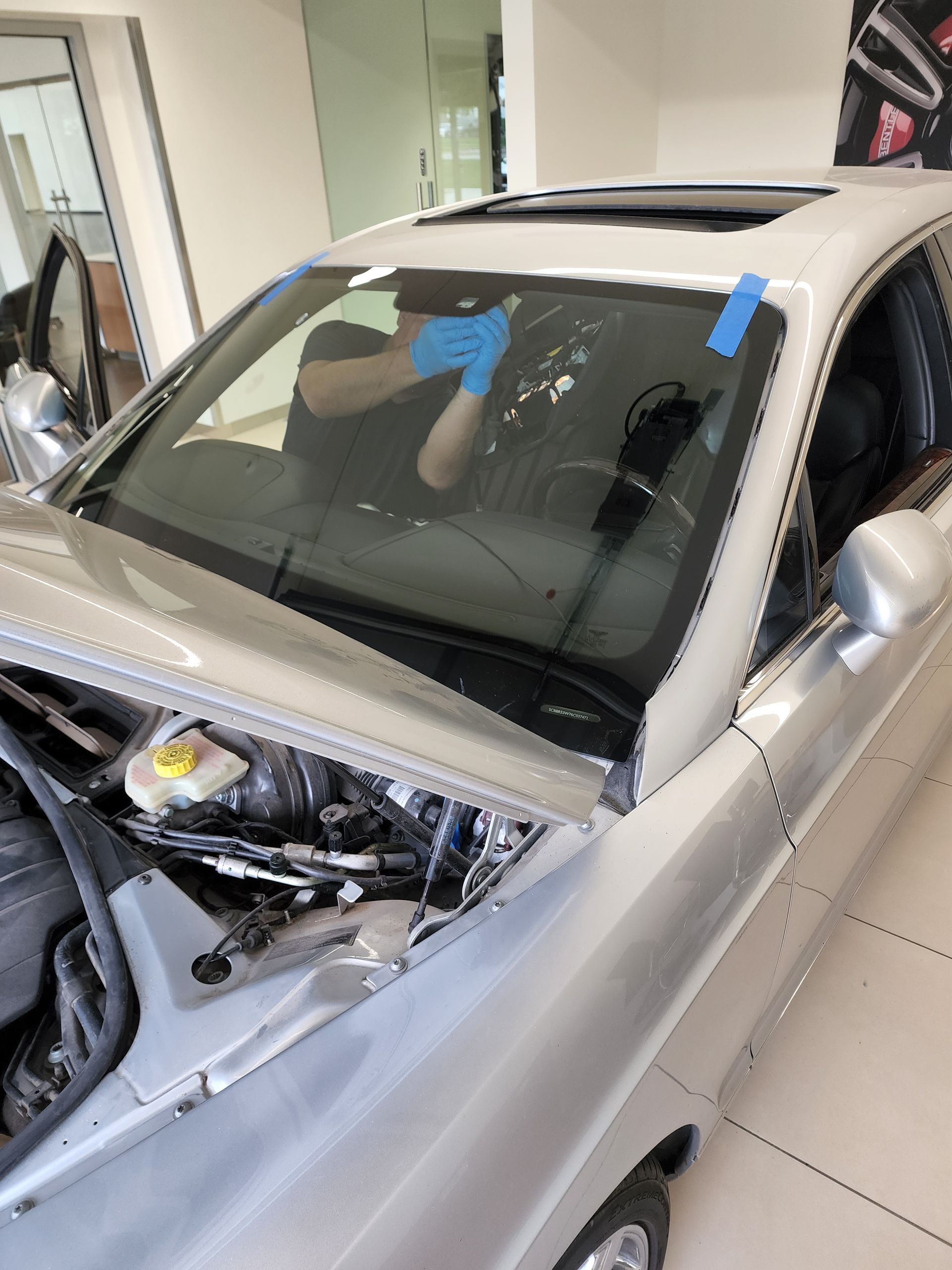 Person in blue gloves working on a silver car's windshield in a bright setting.