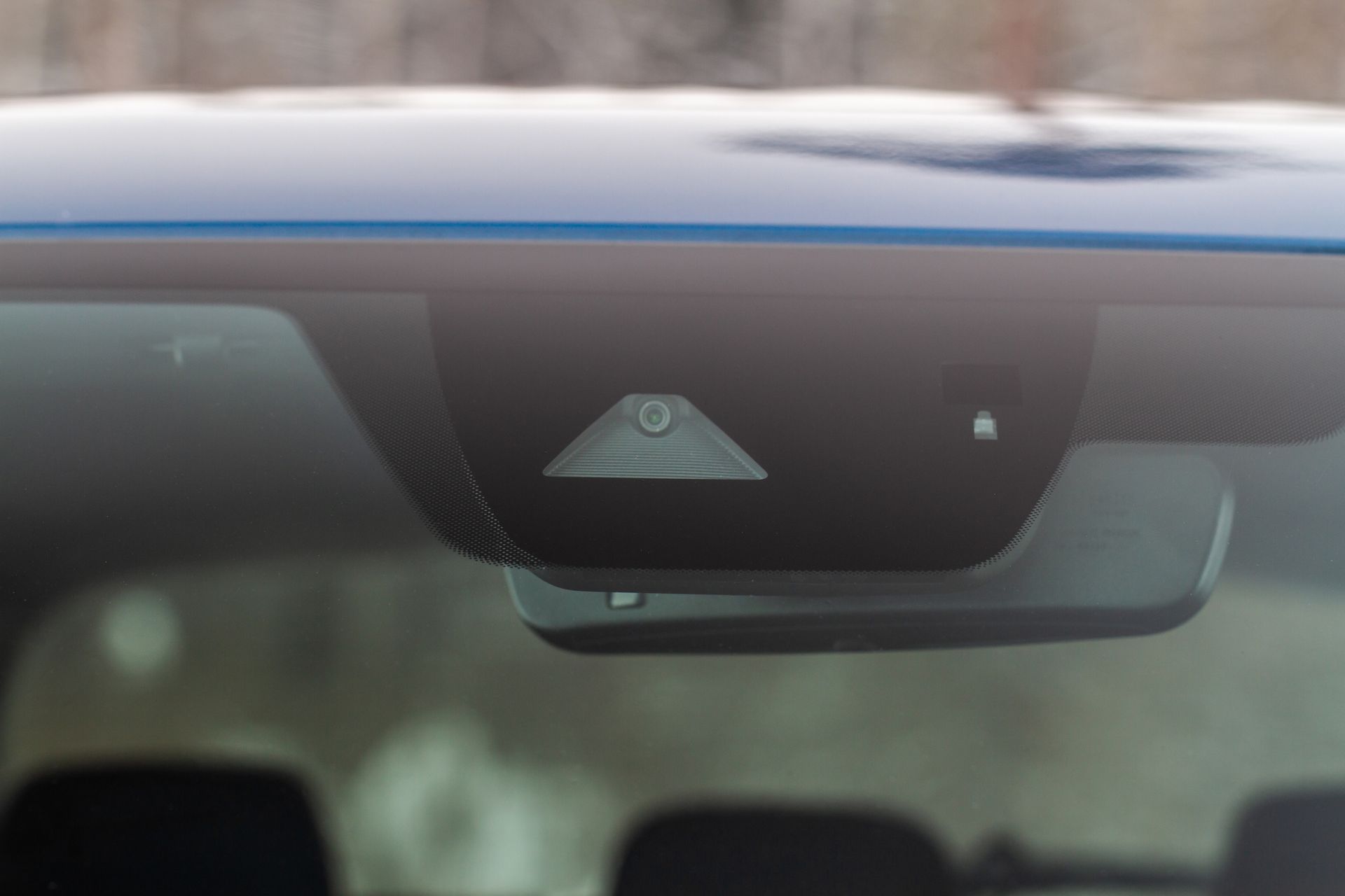 Close-up of car's interior rearview mirror with sensors; against blue car roof and snowy trees.