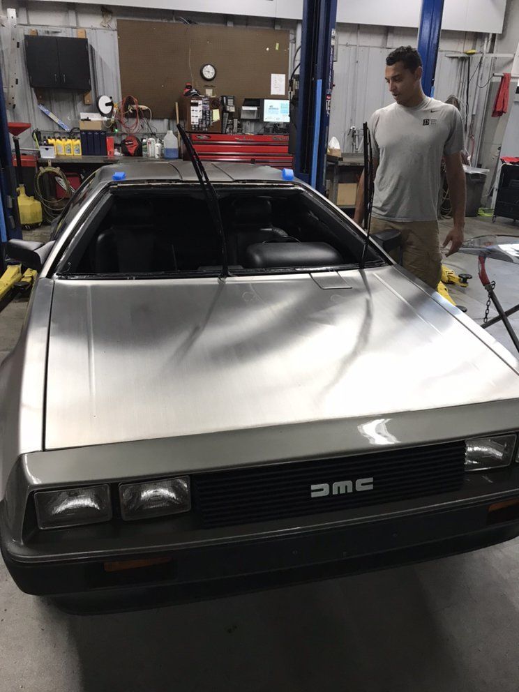 A man inspects a silver DeLorean DMC-12 in a garage. The car has black accents and the 