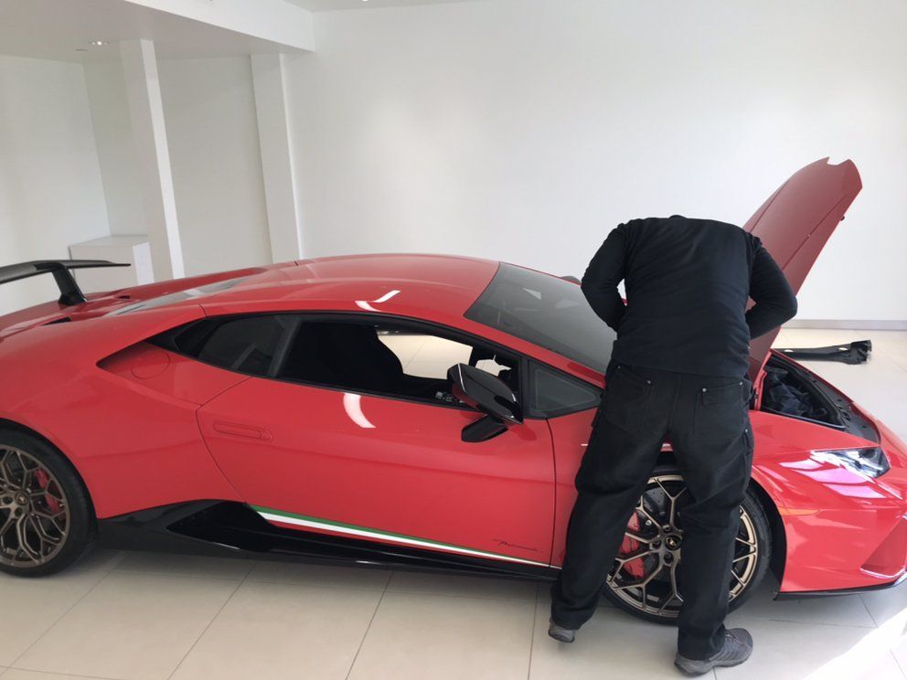 Man in black outfit inspecting the open hood of a red Lamborghini sports car.