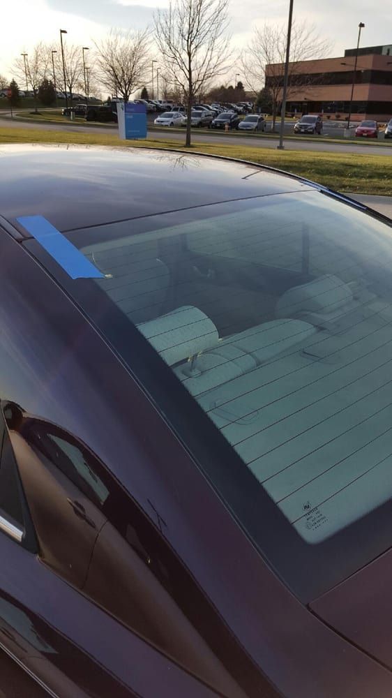 A dark car's back window is taped. The setting is a parking lot with cars and buildings in the background.