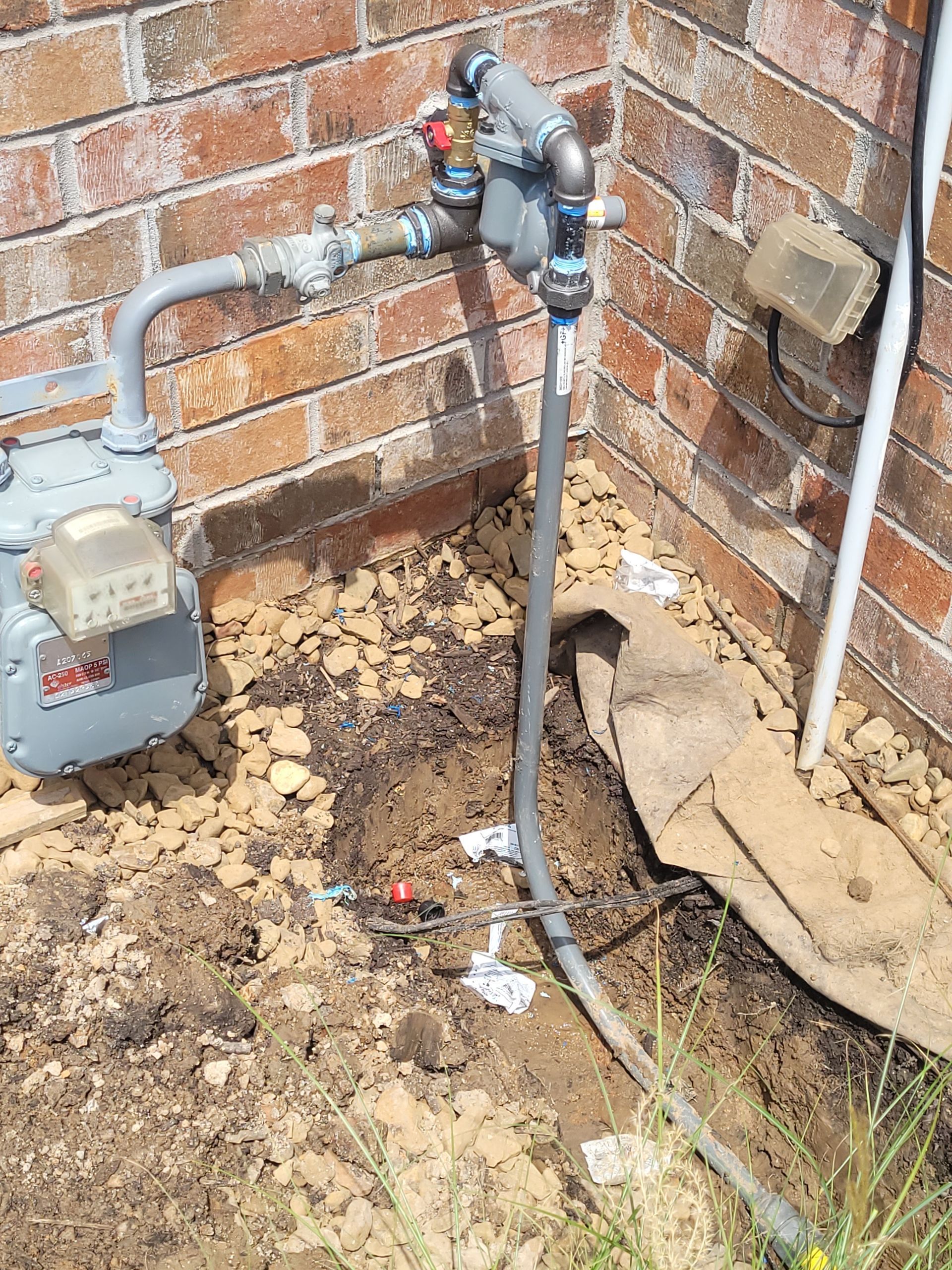 A gas meter is sitting in the dirt next to a brick wall.