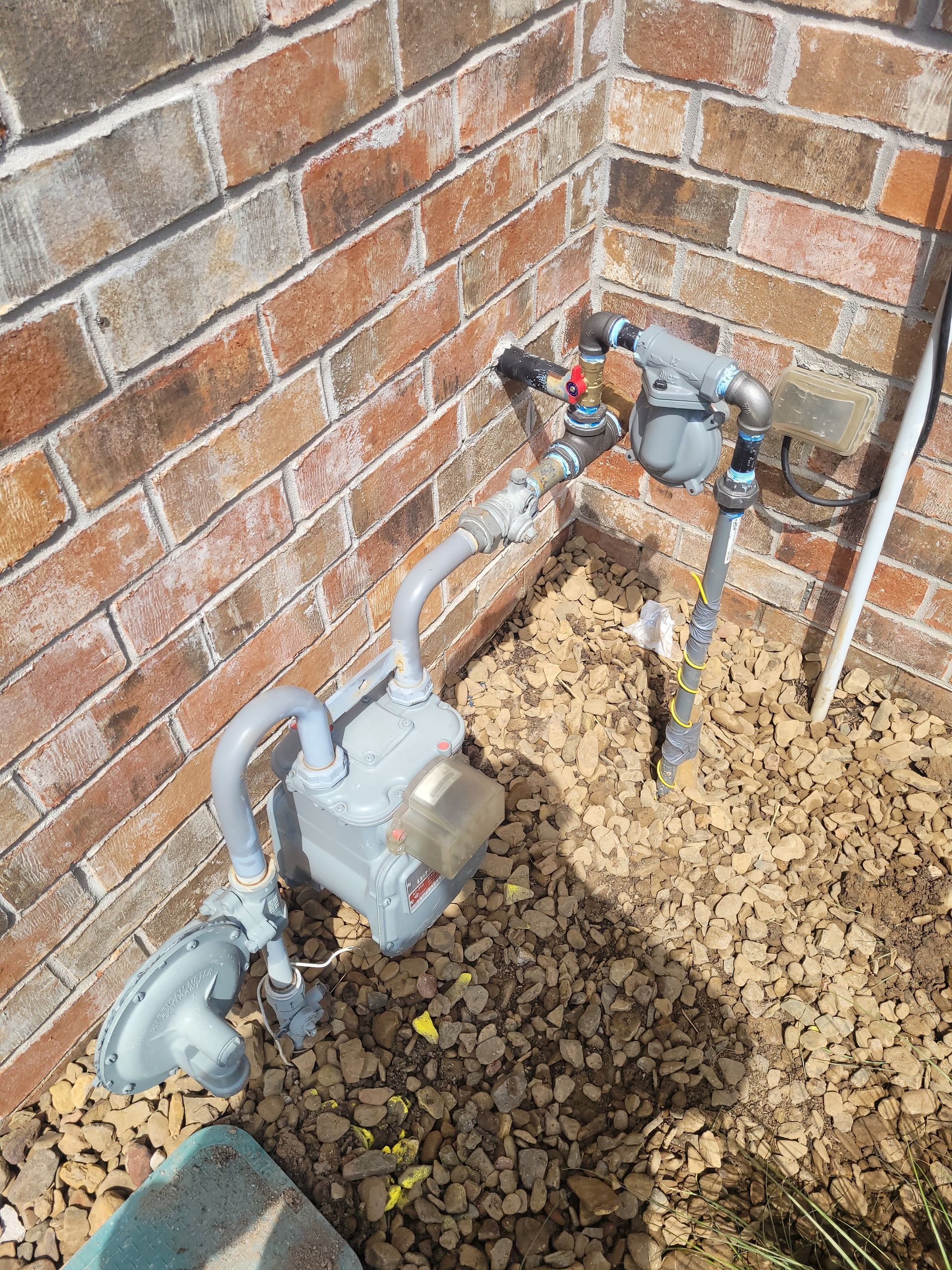 A gas meter is sitting on the ground next to a brick wall.