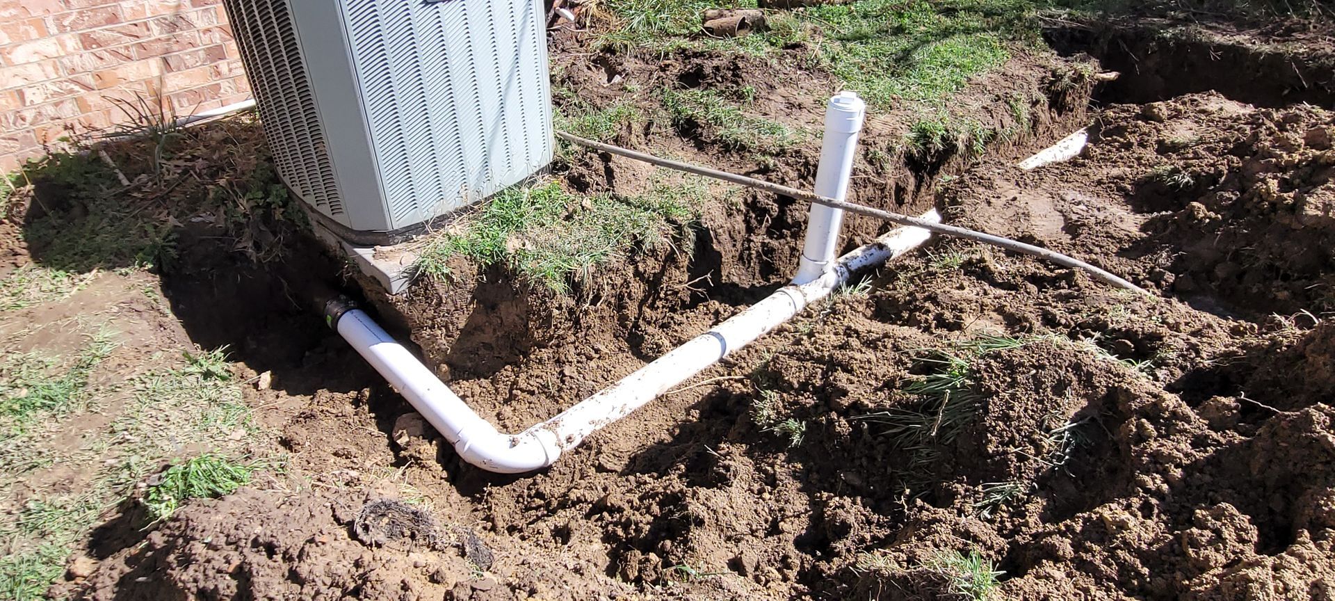 An air conditioner is sitting in the dirt next to a pipe.