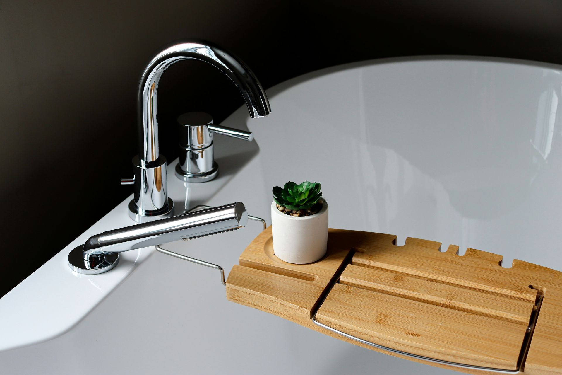 A moody close-up of a shiny white bathtub with chrome faucet supporting a bamboo tray with a tiny potted succulent on top.