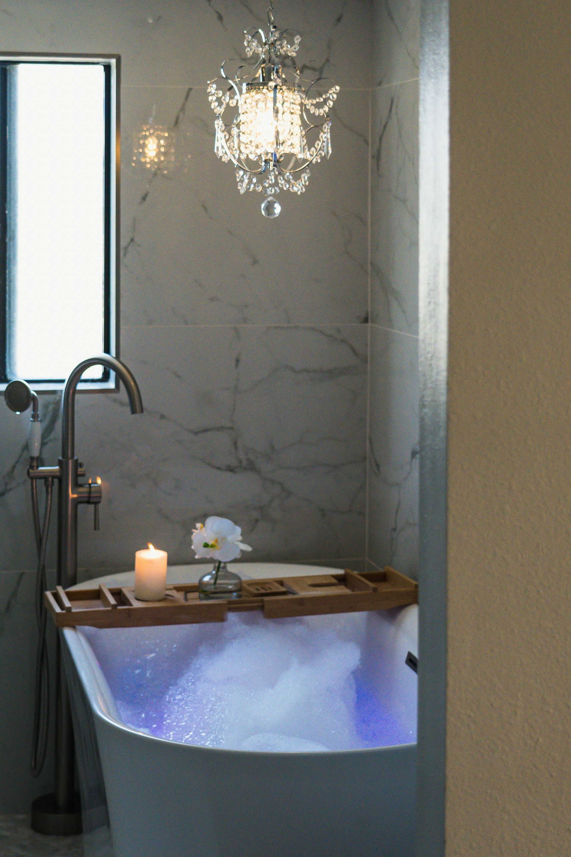 A glamorous free-standing tub surrounded by marble walls and a bright large paned window. A crystal chandelier hangs above.
