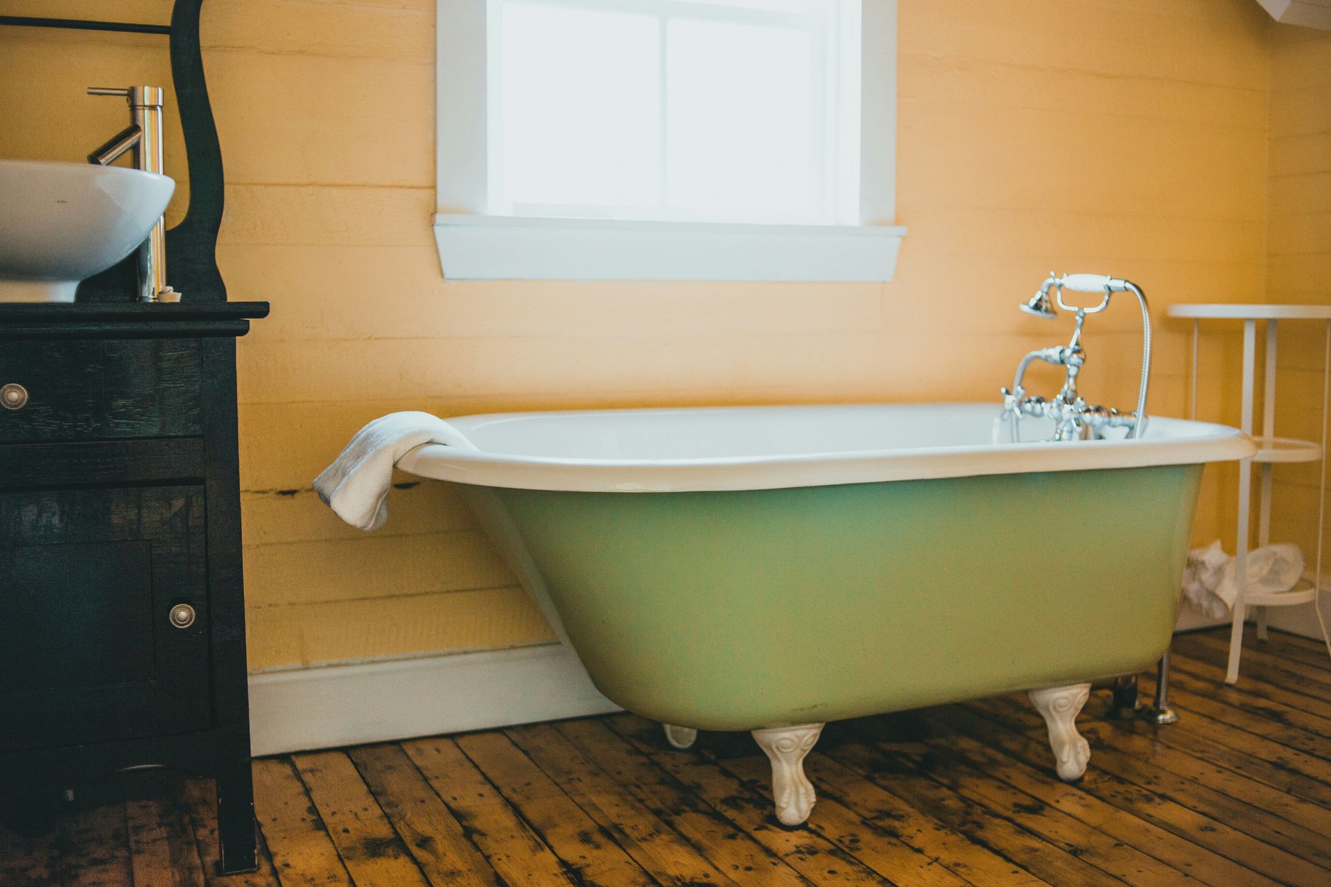 A black wood vanity is next to a bright green cast iron tub. There is a warm wood floor and a bright white window also.