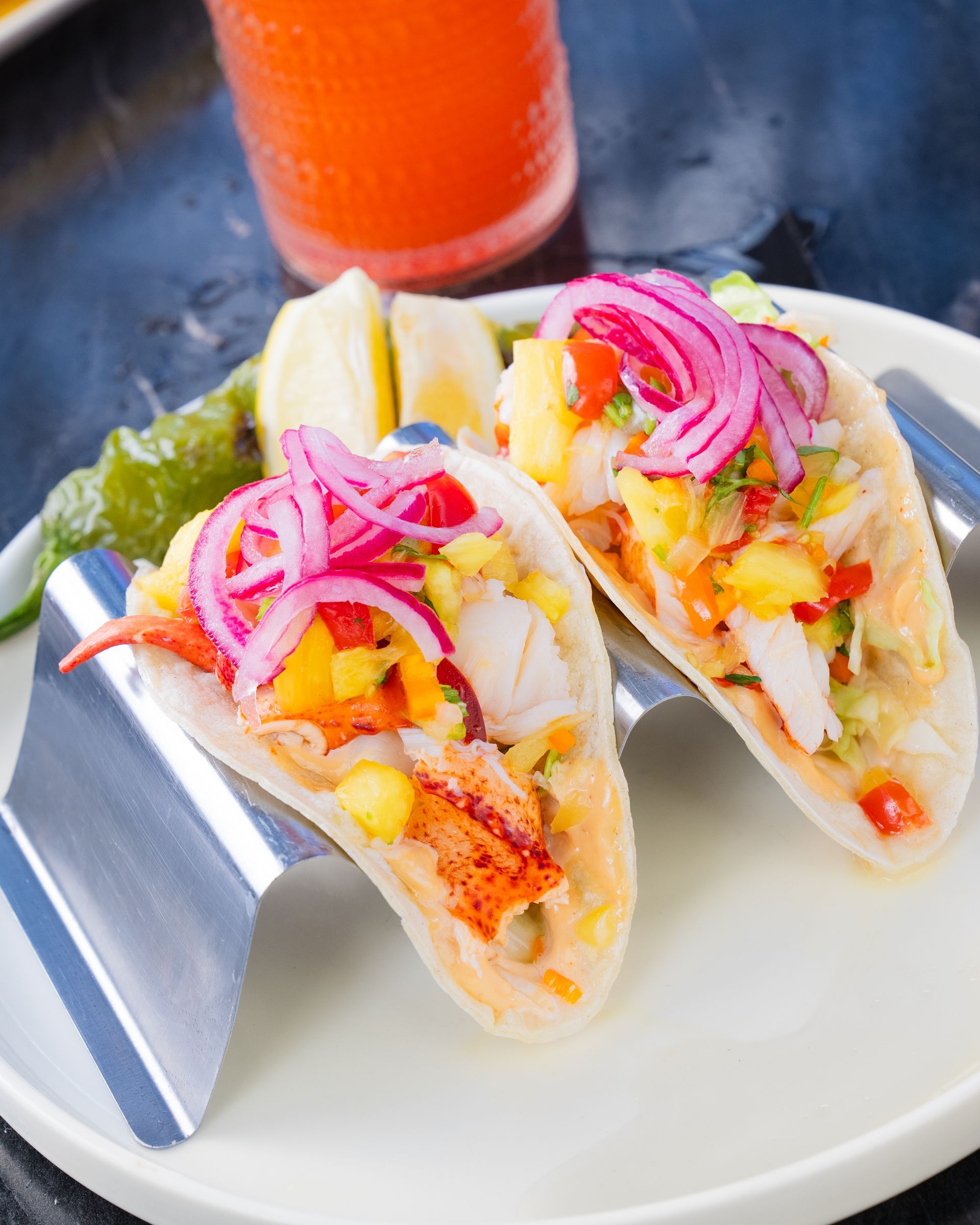 Two lobster tacos with pickled onions and pineapple on a white plate with a drink.