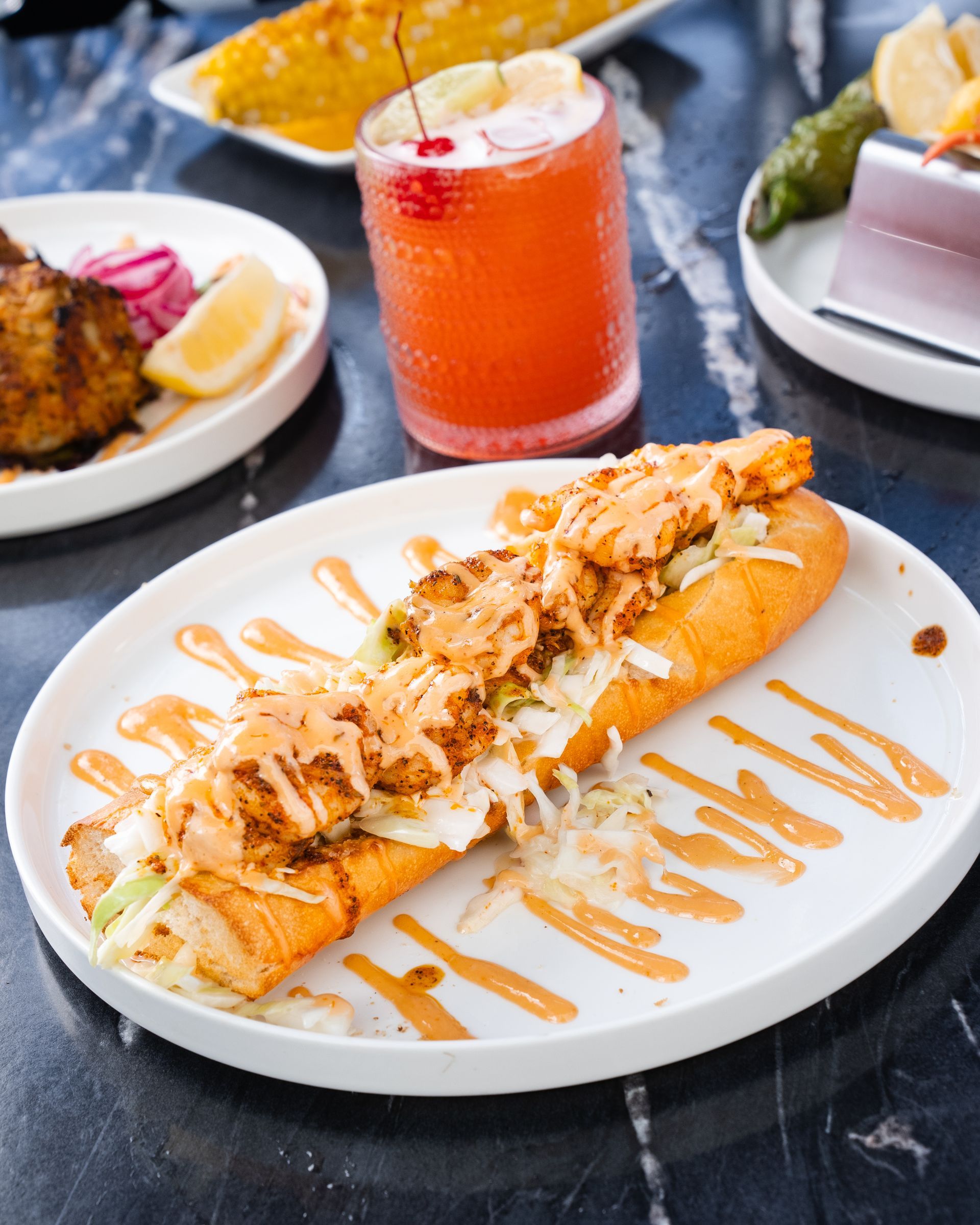 Po' boy sandwich with shrimp and sauce on a white plate, cocktail, and sides on a dark table.
