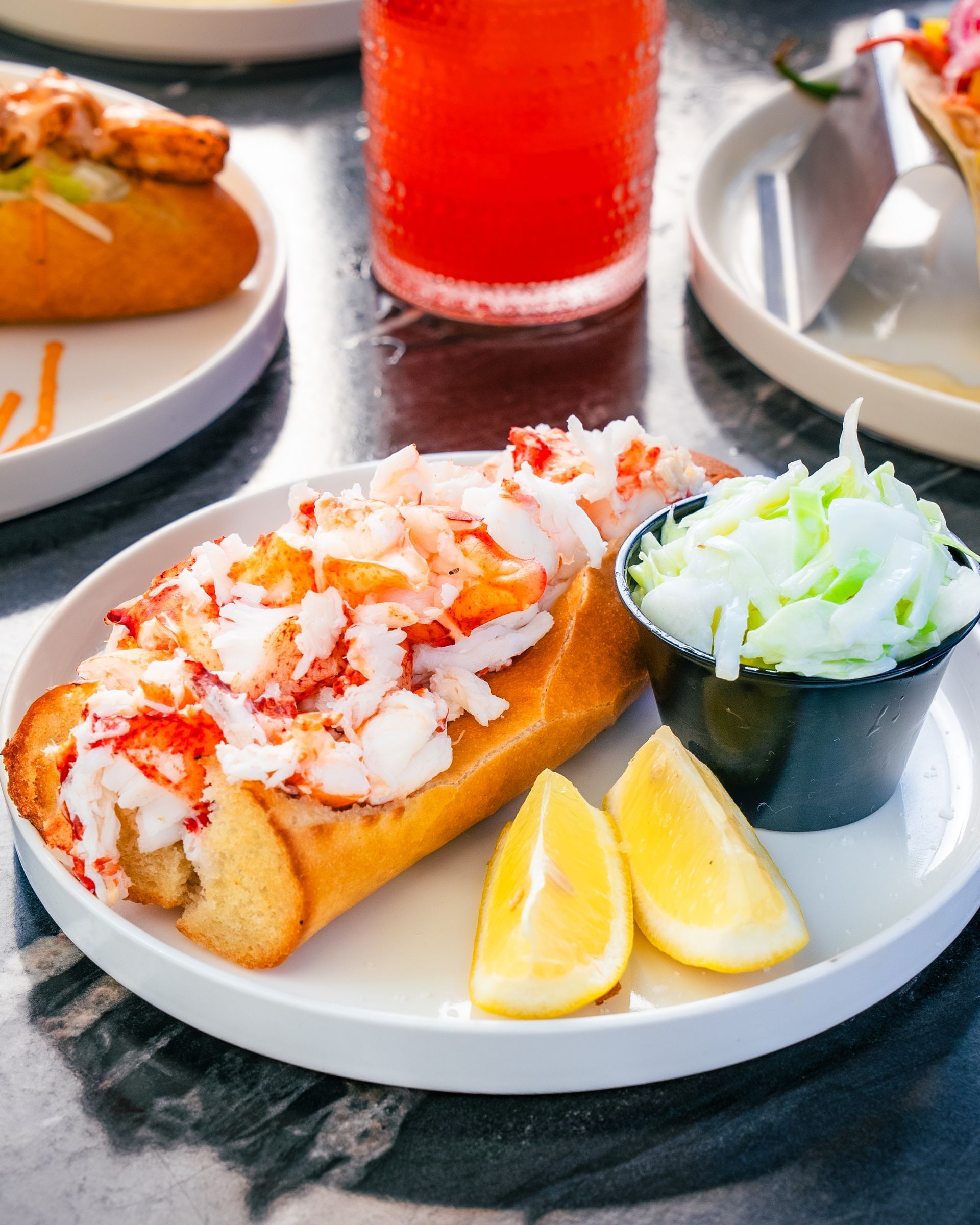 Lobster roll on a white plate, coleslaw, lemon wedges, and a red beverage on a dark table.