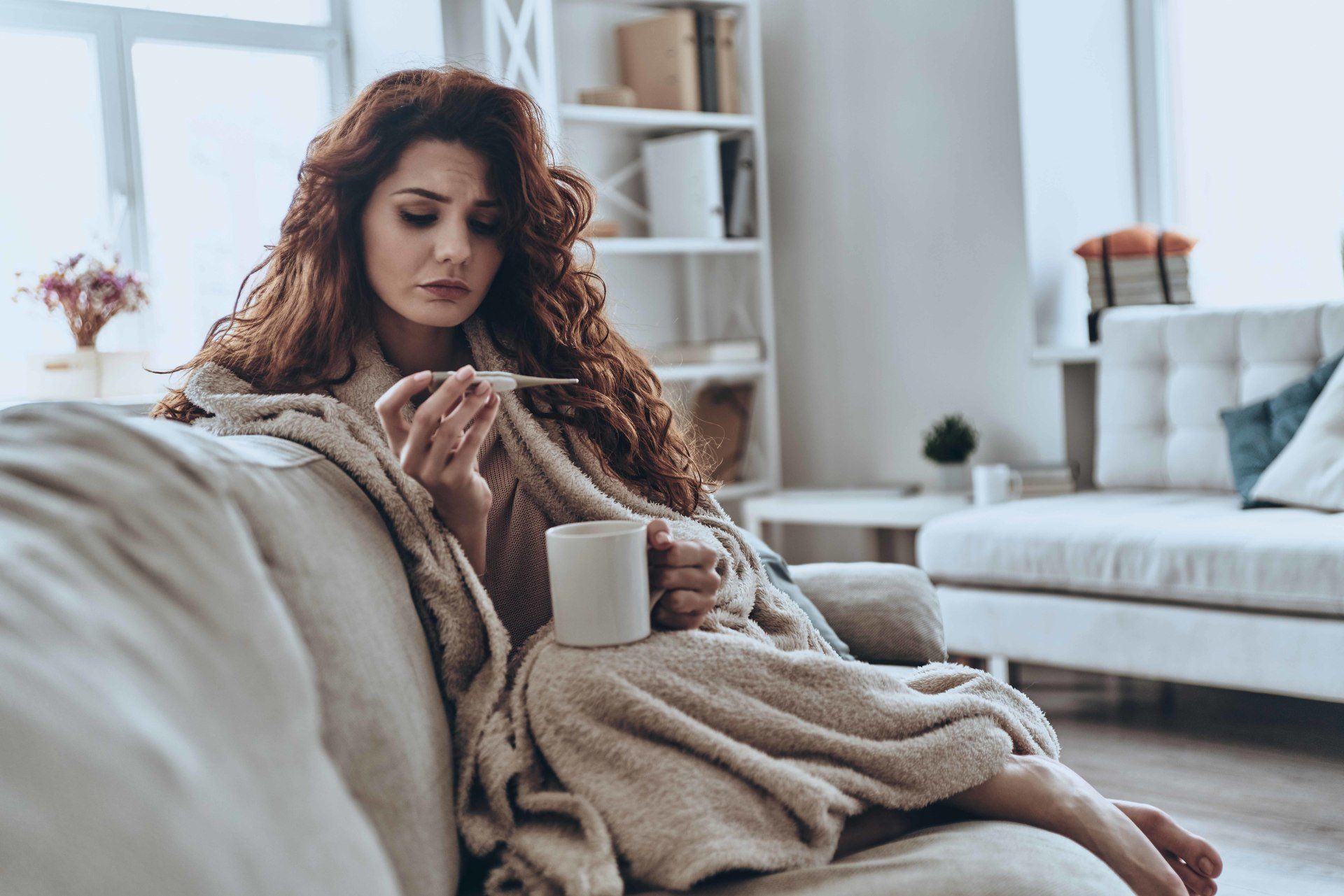 woman sitting on a couch looking at a thermometer and holding tea