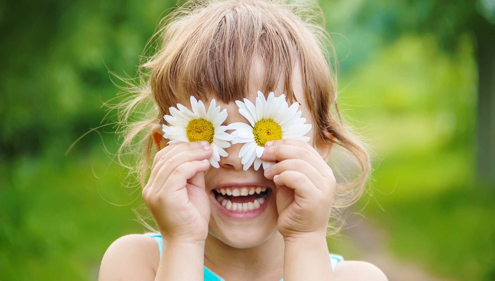 young girl smiling, holding two daisies in front of her eyes