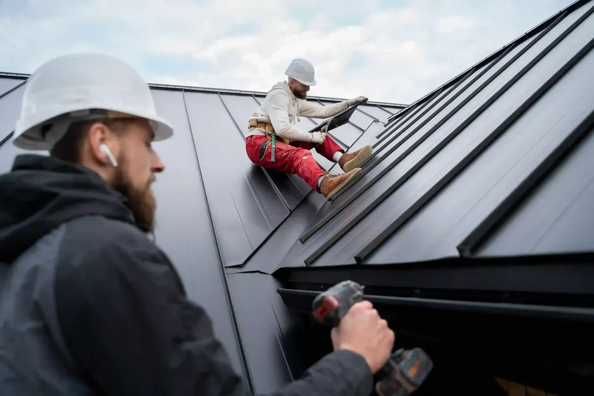 Two roofers working on a black metal roof, one kneeling with a tool in the foreground