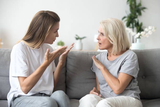 Two women arguing on a gray couch, both gesturing, one younger, one older.