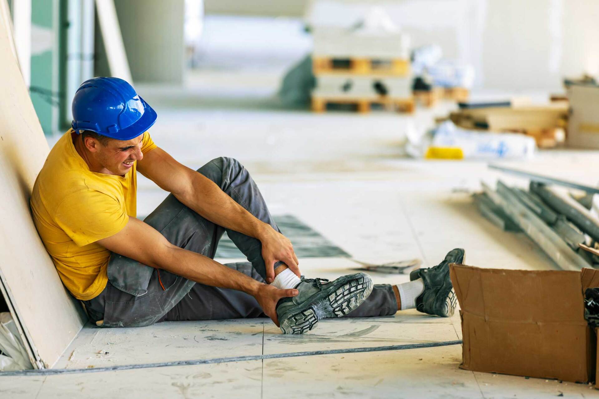 Construction worker on floor holding ankle in pain, blue hard hat, yellow shirt, construction site.