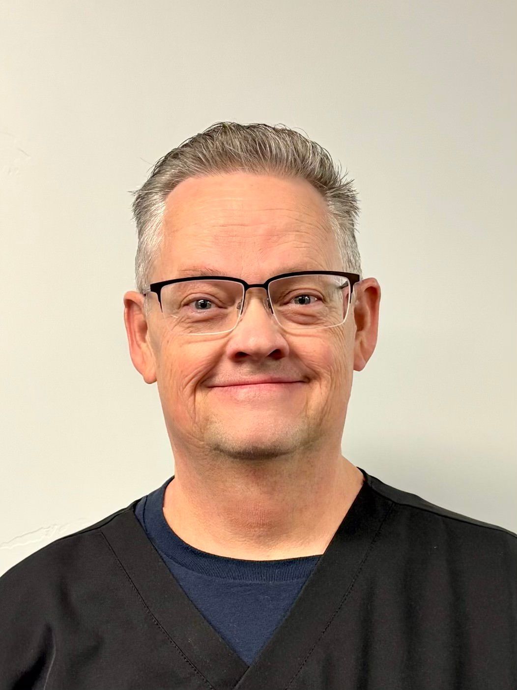 A smiling person with short hair and glasses, wearing a dark blue sweater under a black scrub top, against a light wall.