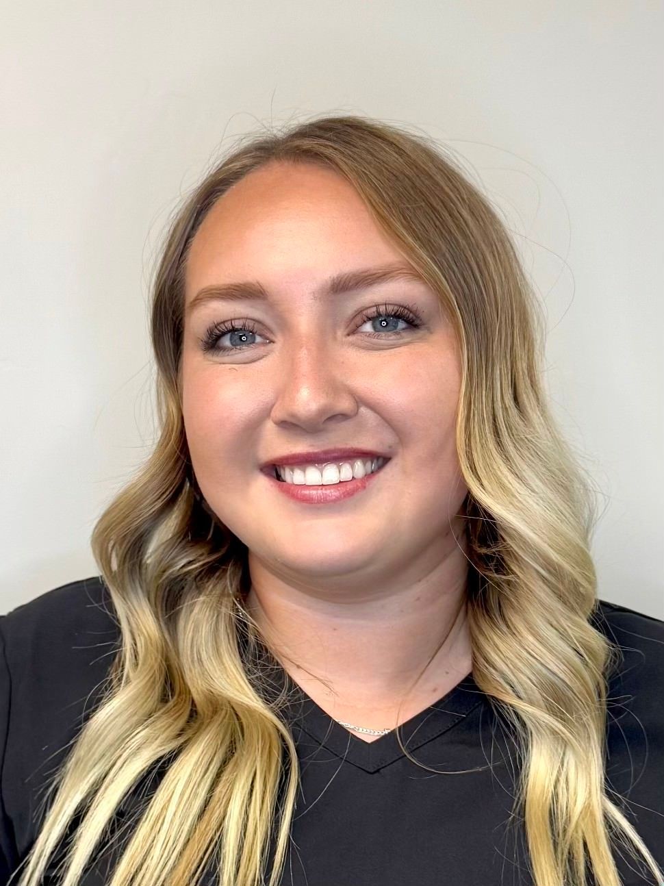 A person with wavy, shoulder-length blonde hair and a V-neck black shirt smiling against a plain, light-colored background.