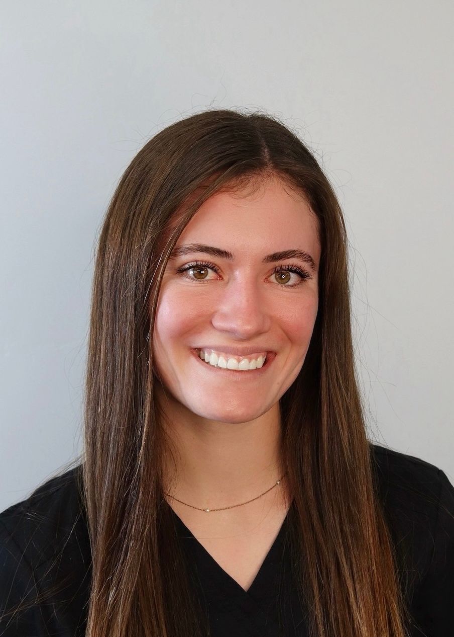A person with long brown hair smiling at the camera, wearing a dark top against a plain light background.