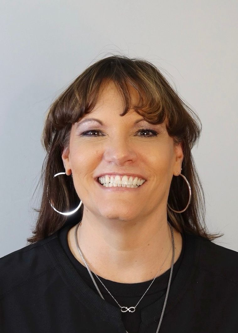 Smiling person with shoulder-length brown hair, large silver hoop earrings, and a black shirt against a plain background.