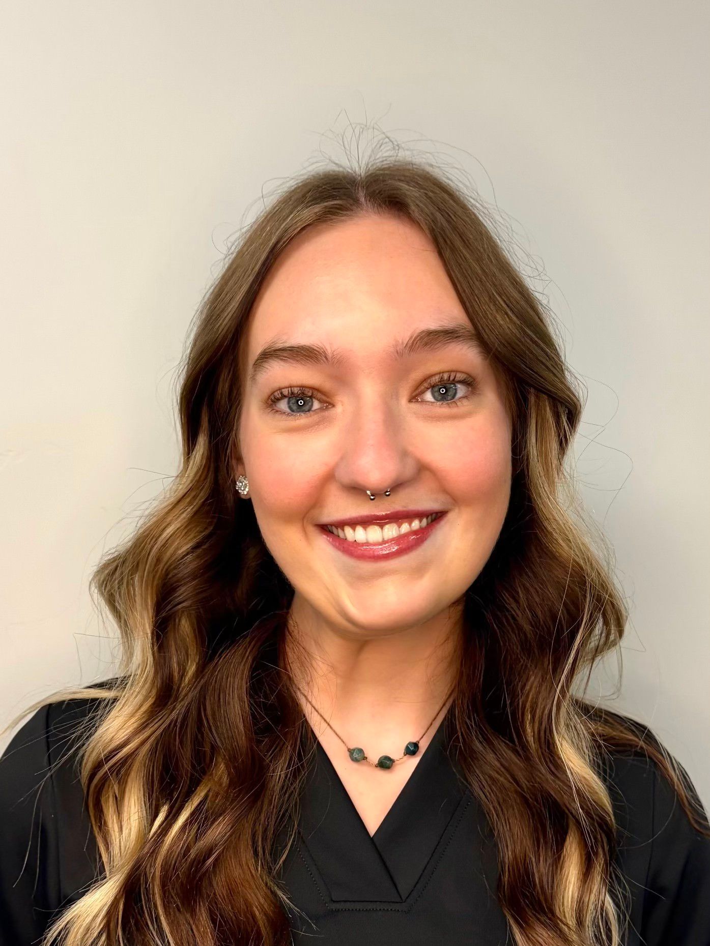 A person with long, wavy brown hair with blonde highlights smiles at the camera, wearing a dark top and a beaded necklace.