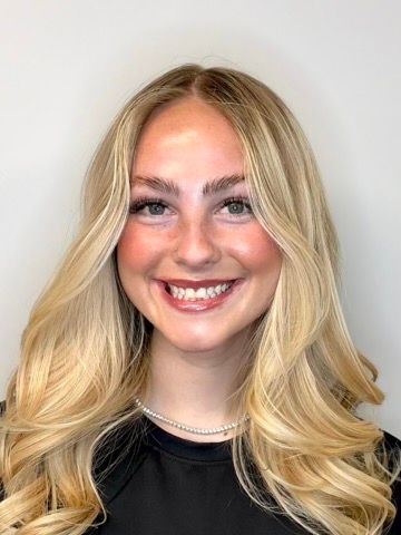 A person with long, blonde, wavy hair and a friendly smile, wearing a black shirt and a silver chain necklace.