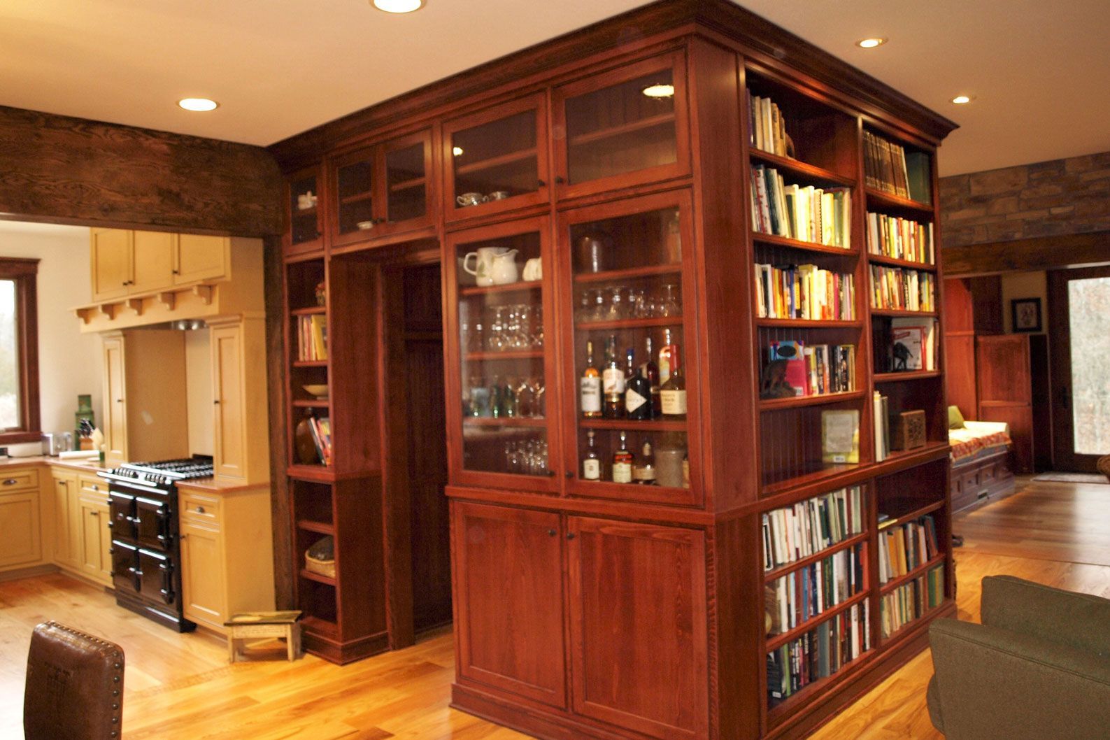 Large wooden bookcase with glass doors and open shelves divides a kitchen and living area.