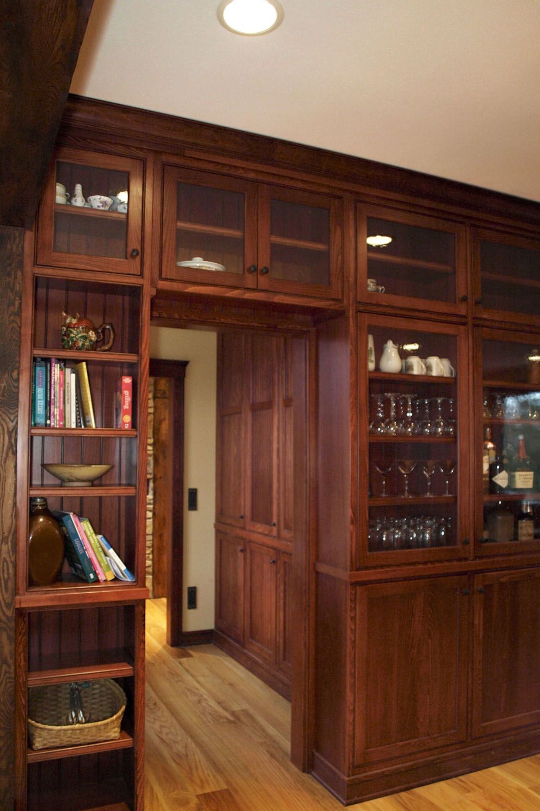 Wooden cabinets with glass doors, bookshelves, and pantry, in a warm-toned hallway with wood flooring.