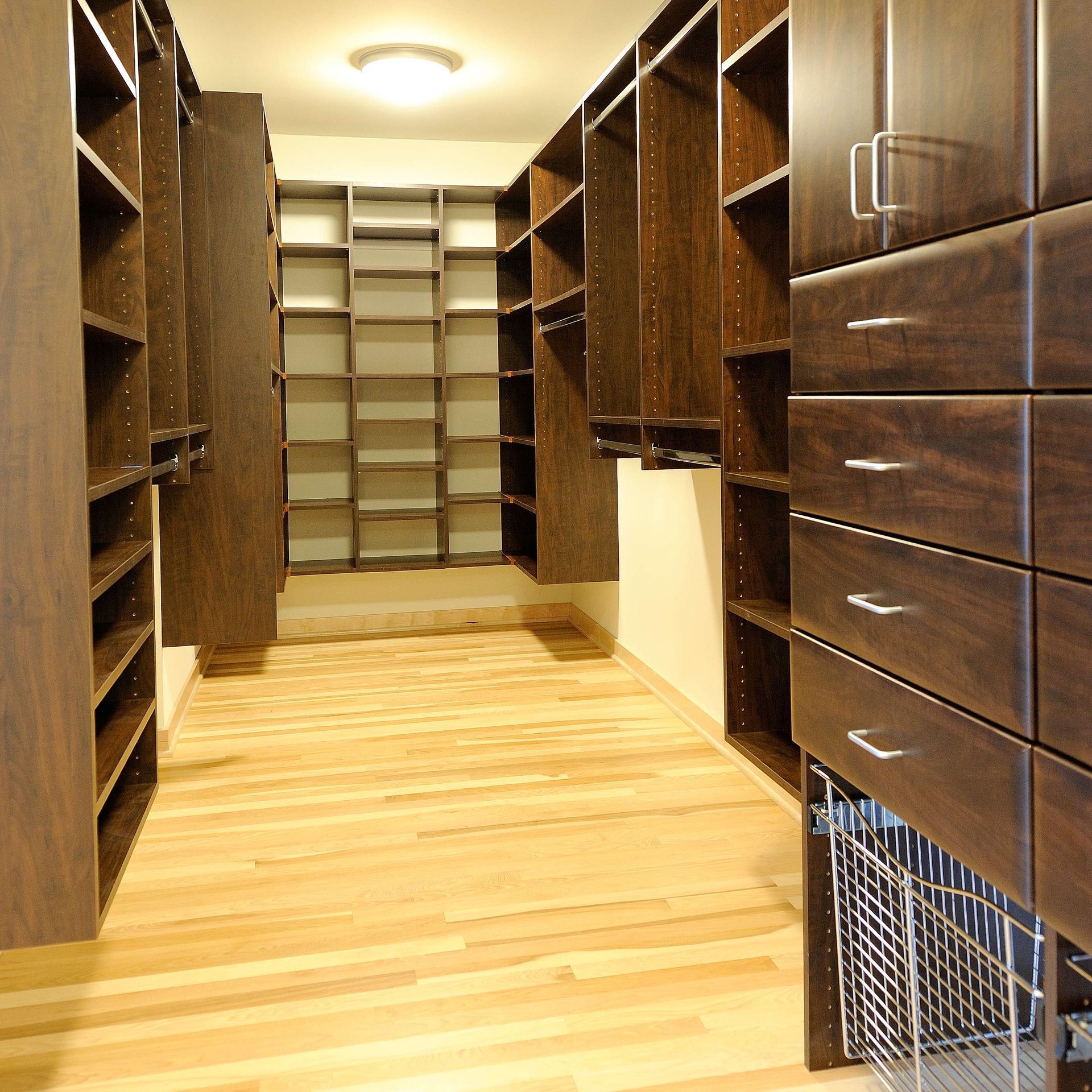 Walk-in closet with dark wood shelves, drawers, and hanging rods. Light wood floor and off-white walls.