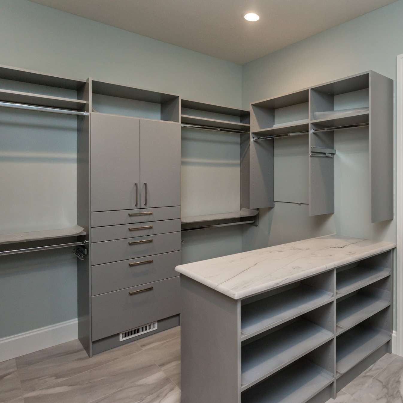 Gray walk-in closet with shelves, hanging rods, and a central cabinet. A light-colored countertop and shelves are in the foreground.