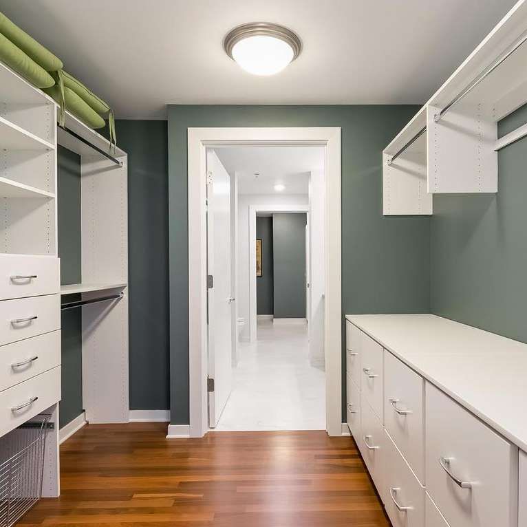 Walk-in closet with white shelves and drawers, green wall, hardwood floor, and open doorway to hallway.