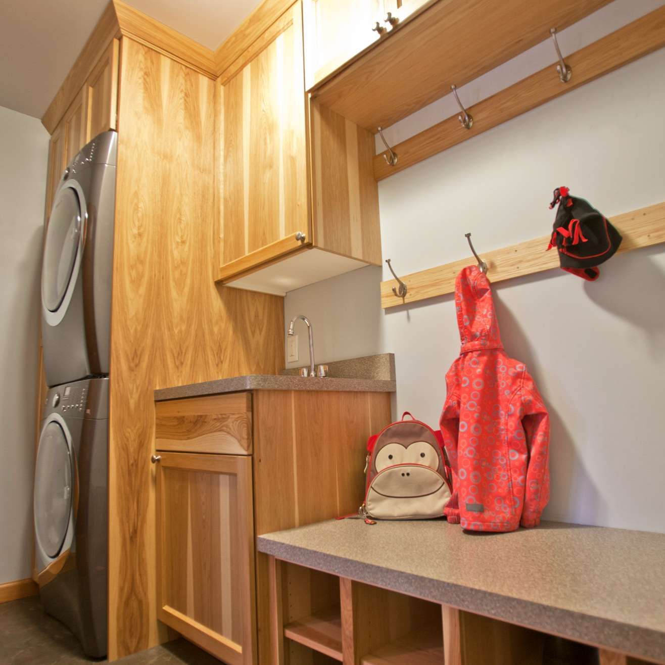 Laundry room with stacked washer/dryer, sink, wooden cabinets, and coat rack with backpack and red jacket.